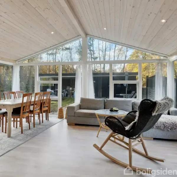 En moderne stue med en spisebord til seks personer står ved siden af en sofa med tæppe. Vinduerne giver udsigt til en have med træer.