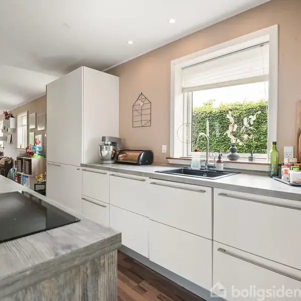 Køkken med hvide skabe og indbygget vask, hvor køkkenredskaber og krydderier er placeret. Et stort vindue giver udsigt til en grøn have. Baggrunden viser en opholdsstue.