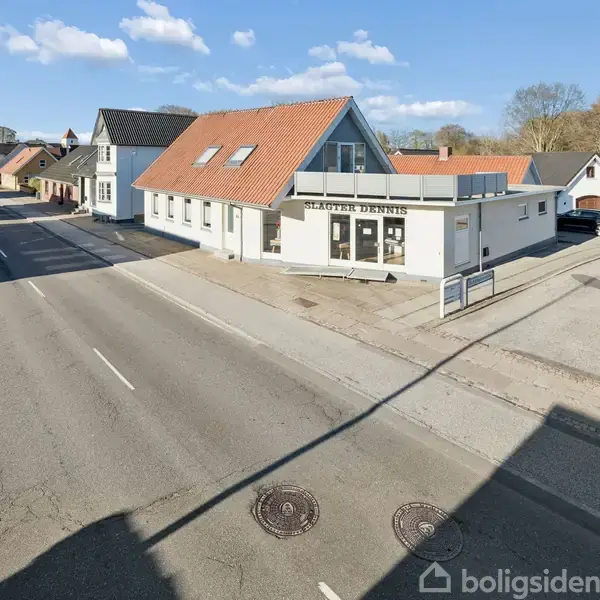 Et hvidt hus med skiltet "Salon Dennis" ses fra gaden. Det står i et bymiljø med flere bygninger og en parkeringsplads på højre side.