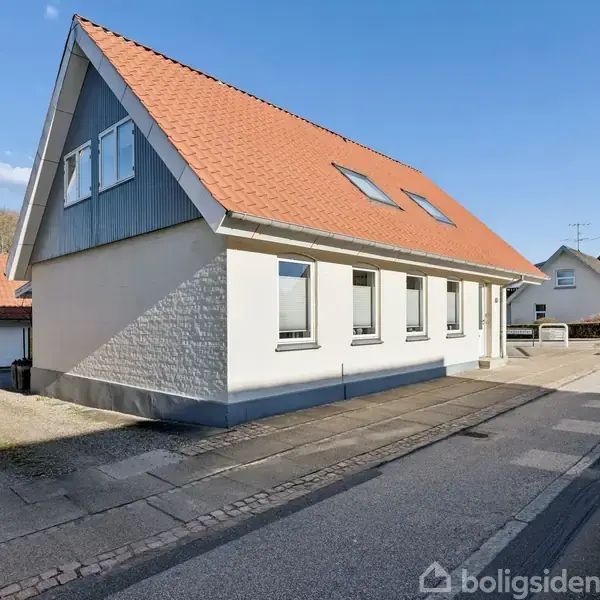 Et hvidt hus med rødt tegltag og flere vinduer står ved en stille vej på en solrig dag. En bilfri gade passerer forbi.