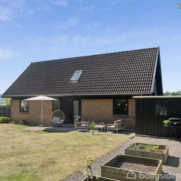 Et brunt murstenshus med et sort tegltag står i en frodig have. Foran huset er der en terrasse med havemøbler og parasol. Der er også hævede plantekasser i haven.