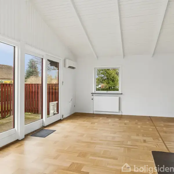 Et lyst stueområde med bræddegulve og en moderne pejs. Store vinduer og glasdør åbner mod en terrasse, omkranset af en rød træhegn. Væggene er hvide, og der er et hvidt loft.