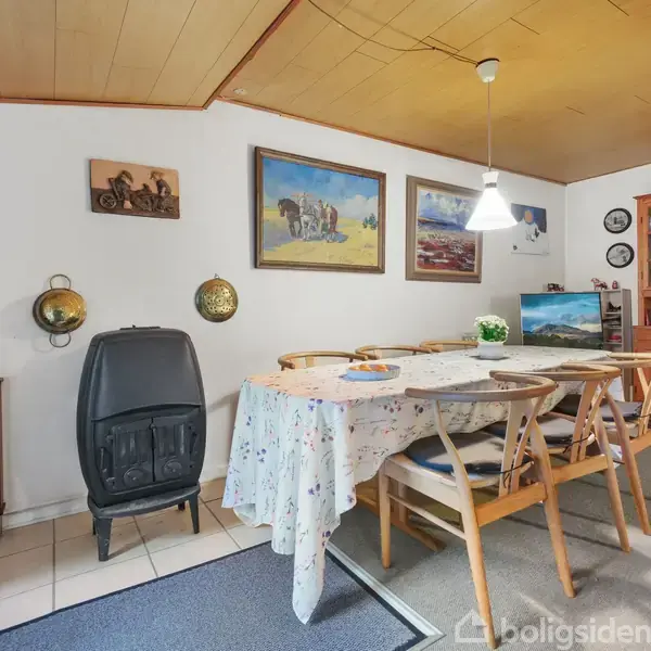 Et spisebord dækket med en blomsterdug er omgivet af træstole i en stue med træloft. En sort brændeovn står ved siden af, og væggen er dekoreret med malerier.