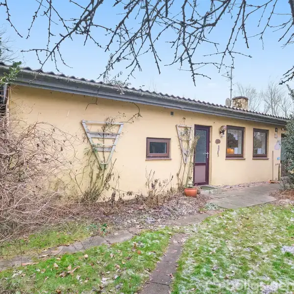 Huset står stille i en have med bar pletvis græs. Væggene er lysegule, og der er flere firkantede vinduer. En dør fører ud til en flisebelagt gangsti.