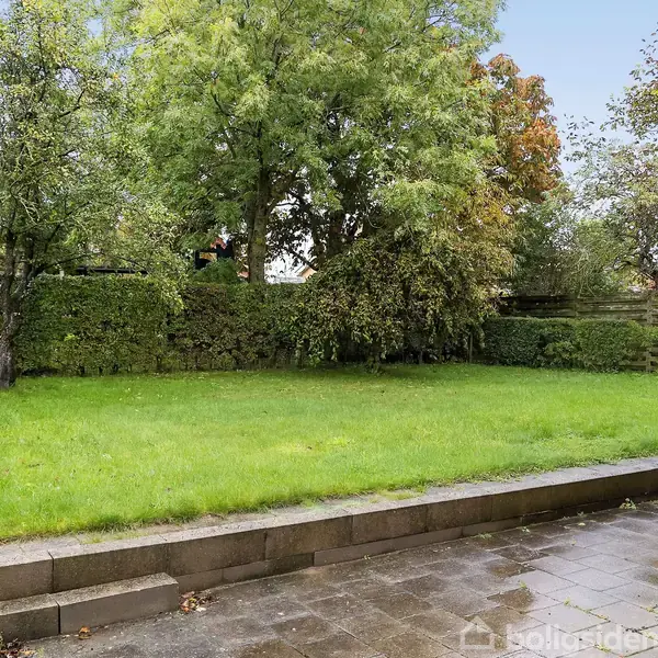 Græsplæne omgivet af hække og træer, der står stille på en regnvåd dag, med en stenterrasse i forgrunden ved siden af en murstensbygning.