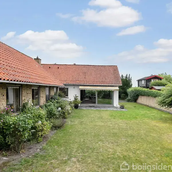 Et enfamiliehus med rødt tegltag og gul murstensfacade står omkranset af en grøn have. Huset har store vinduer, og der er en hæk og en mur i baggrunden.