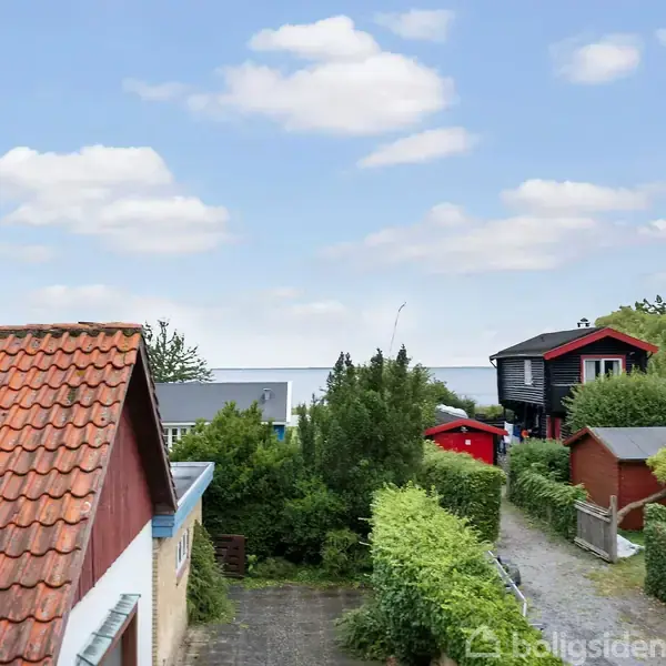 Røde og sorte huse med tage omgivet af grønne buske og træer. En klar blå himmel med få hvide skyer ses i baggrunden.