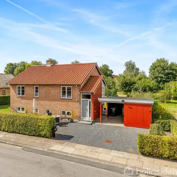 Et rødt murstenshus står stille i et beboelseskvarter med hæk indhegning, grusindkørsel og en rød carport. Omkring er der grønne træer og en blå bil holder på vejen.
