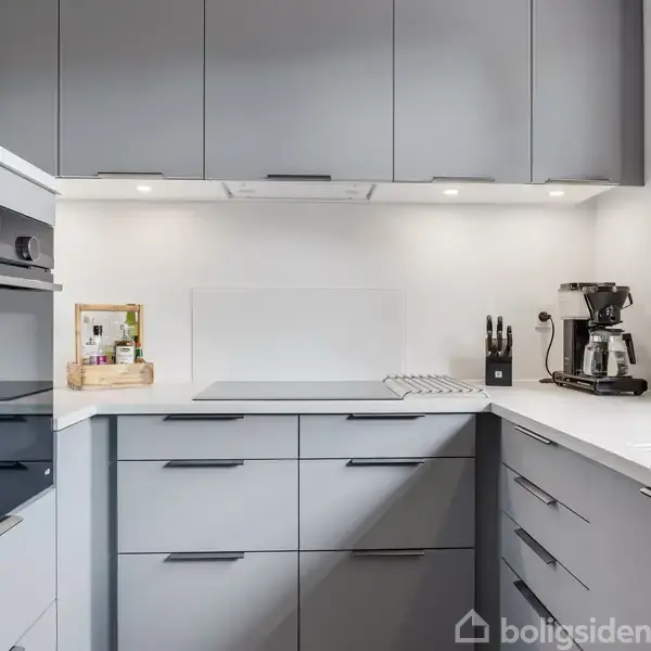 Et moderne køkken med grå skabe er i fokus. En kaffemaskine står på bordpladen ved vasken, mens en ovn er indbygget i væggen. Lyse vægge og god belysning skaber en ren atmosfære.