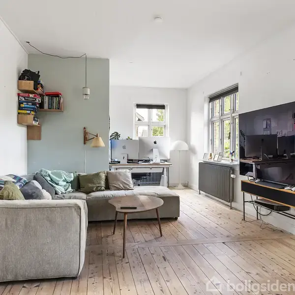 En stue med en grå sofa foran et stort tv, placeret på et bord. Et arbejdsområde med skrivebord og stol ved et vindue. Hylder med bøger hænger på væggen.