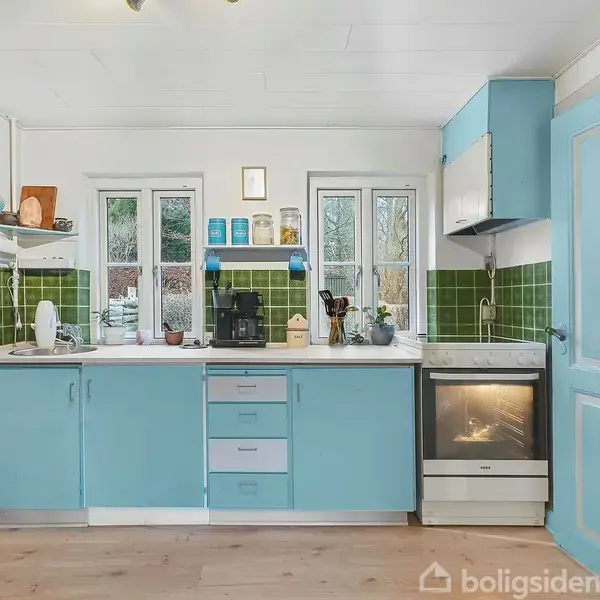 Køkken med lyseblå skabe og skuffer, handling: stilfuldt og ryddeligt, kontekst: omgivet af grønne fliser, lys trægulv og to vinduer med udsigt til udenfor.