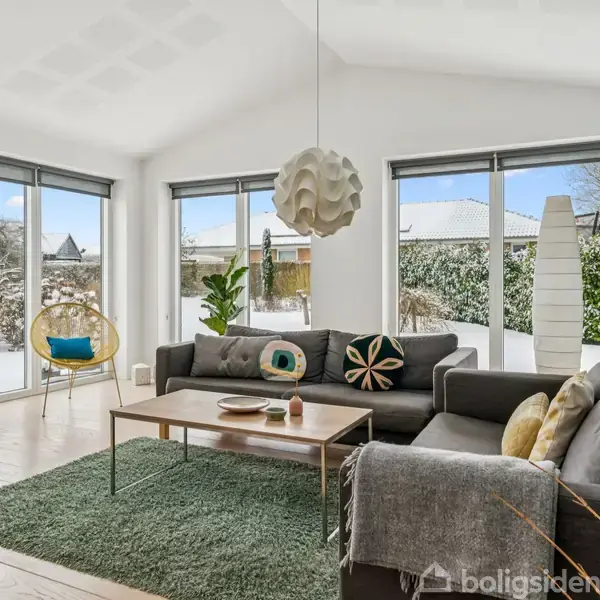 Stue med grå hjørnesofa og sofabord på grønt tæppe. En moderne loftlampe hænger over bordet. Store vinduer viser en snedækket have. Hylder på væggen indeholder bøger og dekorationer.