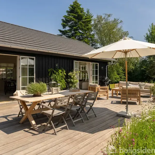 Terrasse med bord og stole under en stor paraply. Ved siden af står en sofa og planter. Baggrunden er en sortmalet bygning med åbne døre og haverige omgivelser.