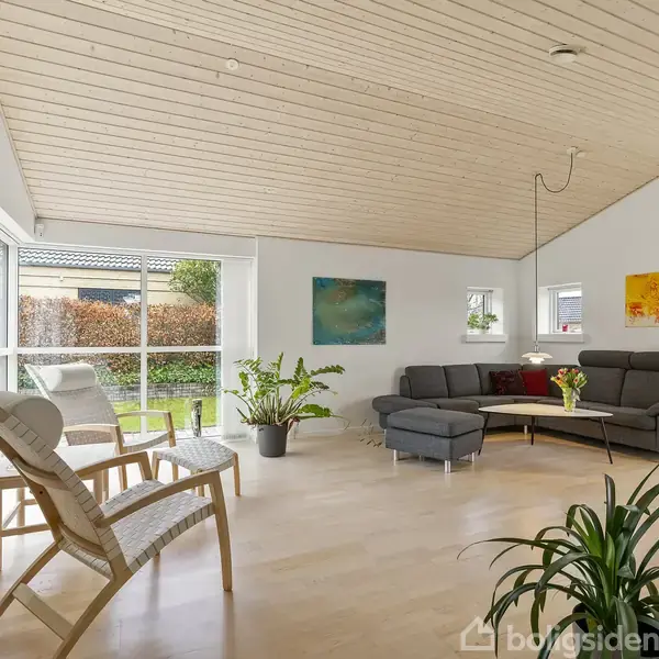 Grå sofa står i en stue med lyst trægulv og hvide vægge. Der hænger farverige malerier på væggene. Sollys strømmer ind gennem store vinduer ved siden af en lænestol. Plante i potte tilføjer grønt.