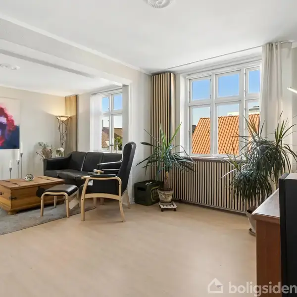 Stue med sort lædersofa og lænestol, der vender mod et sofabord. Store vinduer slipper lys ind. Kunstværk hænger på væggen, og flere grønne planter står i rummet.