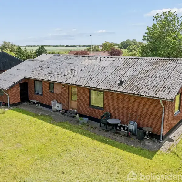 En rød murstenshus med skrånende tag står i en velplejet have. Der er havemøbler ved siden af huset. Omgivelserne består af græsplæne og enkelte træer.