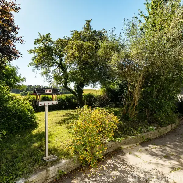 En postkasse står stille ved en have indhegnet af buske og træer, mens solen skinner på en stenlagt terrasse. Markerne ses i baggrunden.