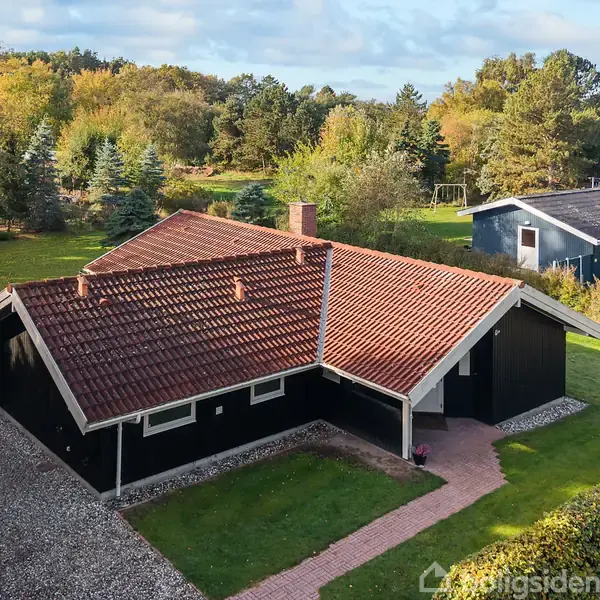 Et sort hus med rødt tag står stille i en grøn have omgivet af træer og en sti. I baggrunden ses et andet hus og skov.