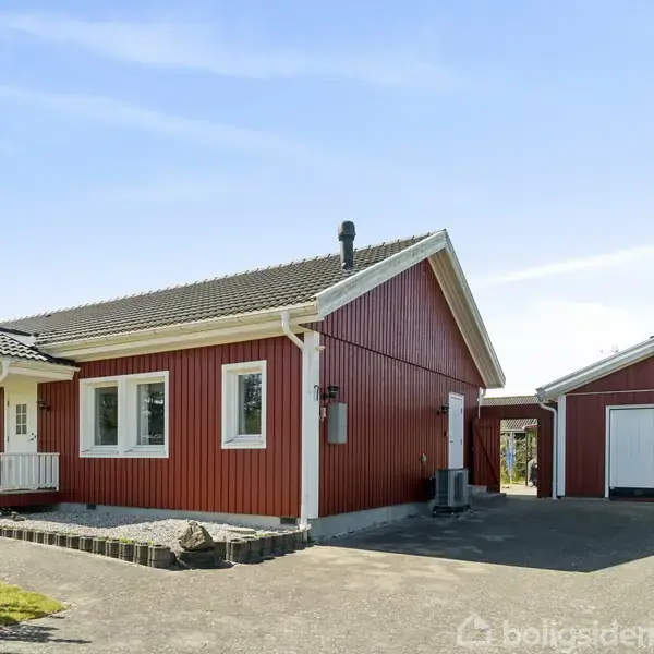 Rødt træhus står stille ved siden af en separat garage. Omgivet af en asfalteret indkørsel og grønne buske under en blå himmel.