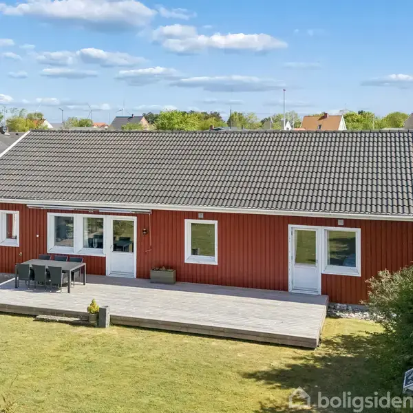 Rødt træhus står stille på en stor træterrasse med havemøbler. I baggrunden ses en grøn have og andre huse i et beboelsesområde under en blå himmel med spredte skyer.