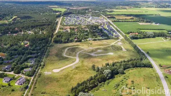 Et åbent grønt område med snoede stier strækker sig gennem et landskab. Omkringliggende skove og marker omkranser et beboelsesområde. Billedet er taget fra en luftperspektiv.