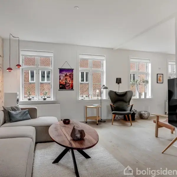 En stue med en stor beige sofa står foran et ovalt træbord. Rummet har to store vinduer, en murstensvæg, kunstværker og en lænestol med lampe.
