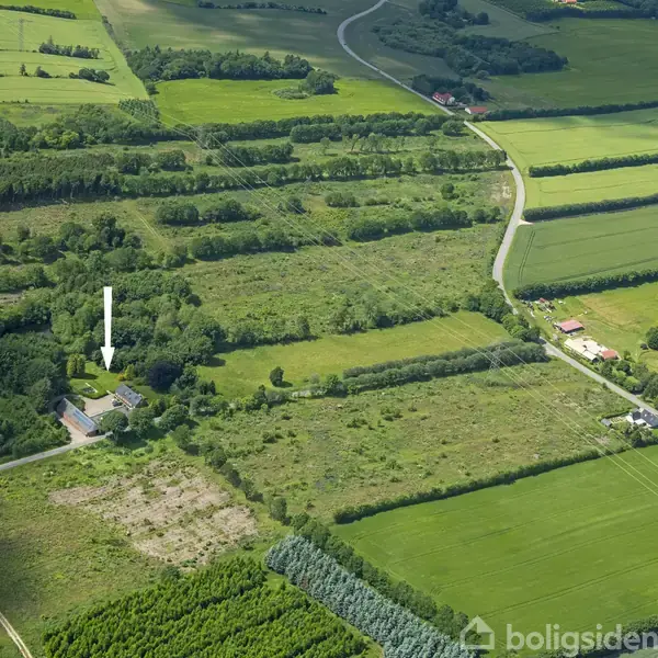Luftfoto af landbrugsområde med marker og skove. En hvid pil peger på en ejendom med bygninger omgivet af træer. Omkringliggende marker adskilles af hegn og veje.