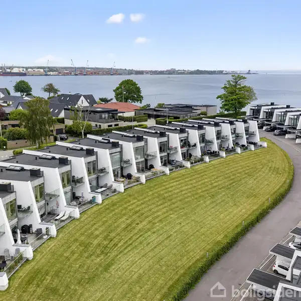 Rækkehuse med hvide, moderne facader står i en kurve langs en vej, omgivet af velplejede græsarealer, med udsigt over en havbugt og en by i horisonten.