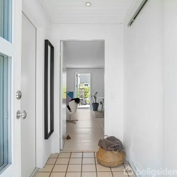 En lys entré med flisegulv fører ind til et rum med store vinduer. En kurv står på gulvet, og et spejl hænger på væggen.