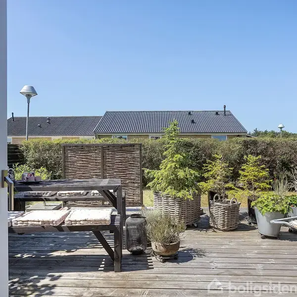 Terrasse med træbord og stole omgivet af kurveplanter. Solrig have med hæk og nabohuse i baggrunden. Klar blå himmel uden skyer.