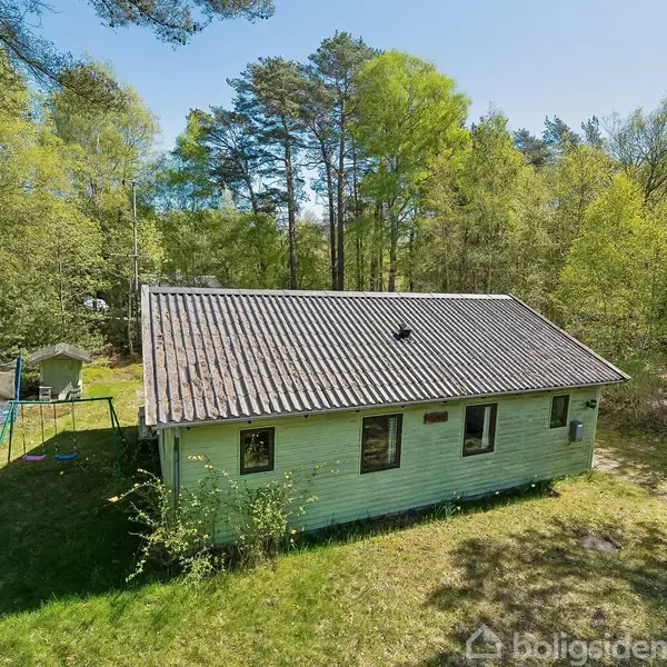 Et grønt træhus står stille omgivet af skovklædt natur. En trampolin ses til venstre i haven. Sollys filtrerer gennem træernes løv, som skaber en fredfyldt atmosfære.