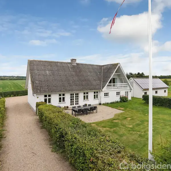 Et hvidt, klassisk hus med sort tag står i en landejendom med grus indkørsel, omgivet af grønne marker og hække. En flagstang med flag er synlig i forgrunden.