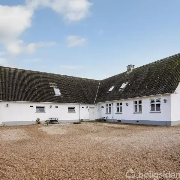 Et hvidt, vinkelformet hus med et sort skråtag står på en grusbelagt gårdsplads. Til højre ses et dansk flag på en flagstang. Himlen er delvist skyet.