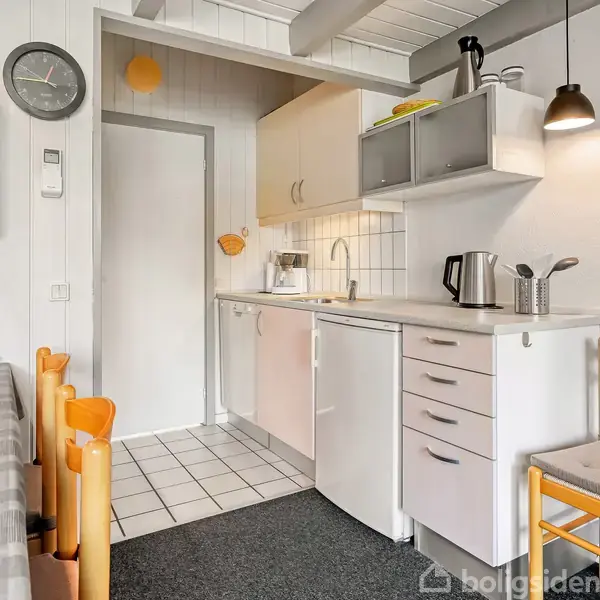 Et kompakt køkken med hvidt køkkenbord og skabe. Kedlen og brødristeren står på bordet. En stol står ved siden af. Uret hænger på væggen. Bordet har ternet dug.