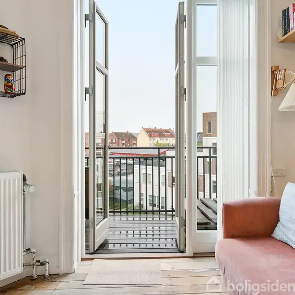 En åben altandør med udsigt til bygninger omgivet af neutrale vægge. En rosa sofa med puder står ved siden af en reol og væglampe. Et radiator ses også.