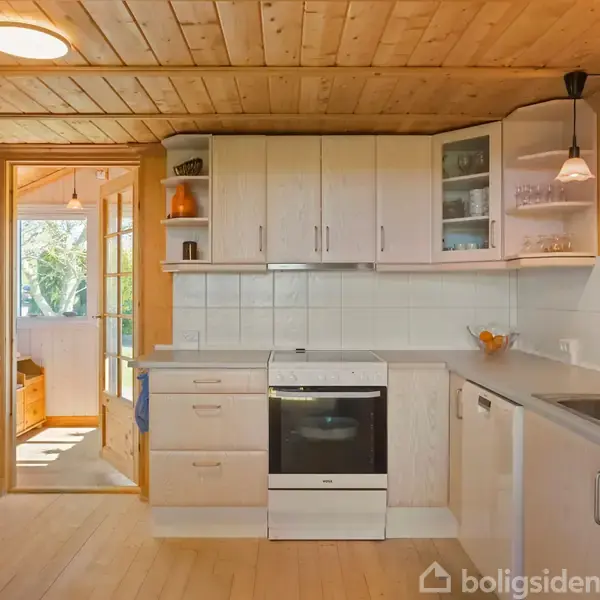 Køkken med hvide skabe og en ovn i midten, trævægge og -loft. Til højre er en lampe over en håndvask, med døråbning til et lyst rum. En fransk dør fører udenfor.