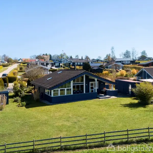 Et hus ligger på en stor, grøn græsplæne med en trægærde i forgrunden. Omkring huset er der flere andre boliger i et boligområde. Himmel er blå og klar.