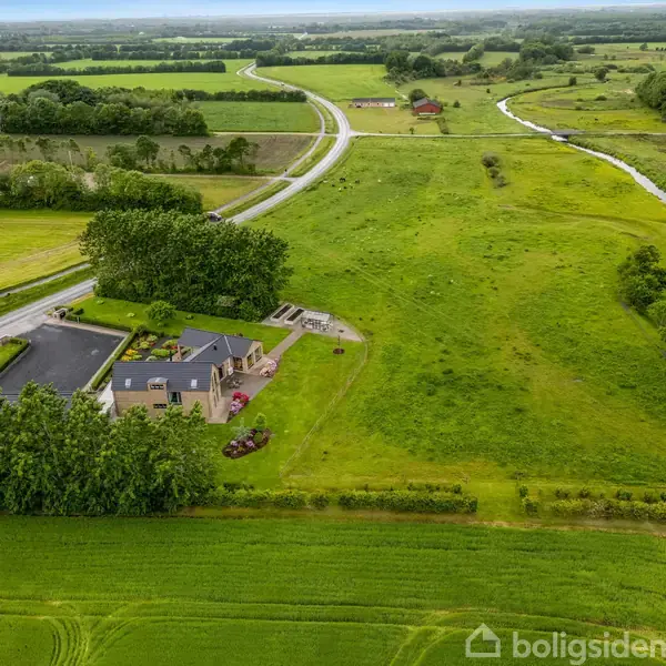 En moderne bolig ligger ved en landevej omgivet af grønne marker og en snoet flod. Flere træer afgrænser ejendommen og markerne.