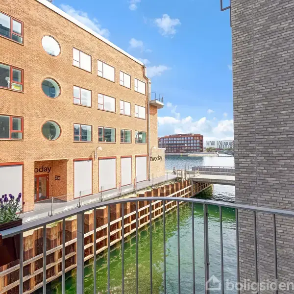 Bygning står ved en kanal med grønligt vand, hvor blomster gror i forgrunden. Murstensfacade har firkantede og runde vinduer. En bro og flere bygninger ses i baggrunden.