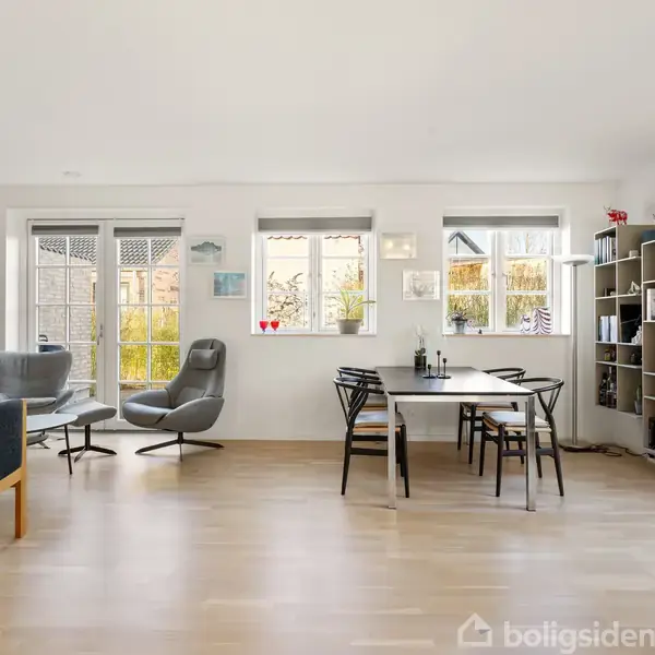 En stue med lyse trægulve har et spisebord med stole til højre og to lænestole til venstre foran vinduer. En reol med bøger og dekorationer står ved siden af bordet.