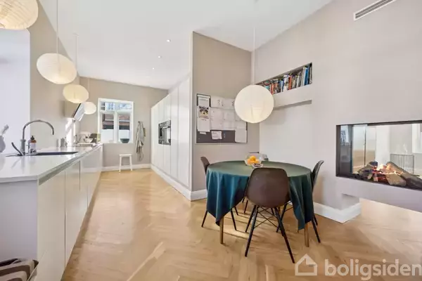 Køkken-alrum med moderne design, hvor et rundt bord med blå dug står foran en pejs. Til venstre er et hvidt køkken med lysopsætning og trægulv. En opslagstavle hænger over bordet.