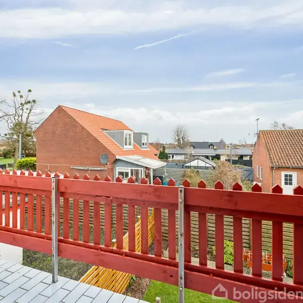 Rød træterrasse omkranser udsigt over nabolag med murede huse, en blå himmel i baggrunden.