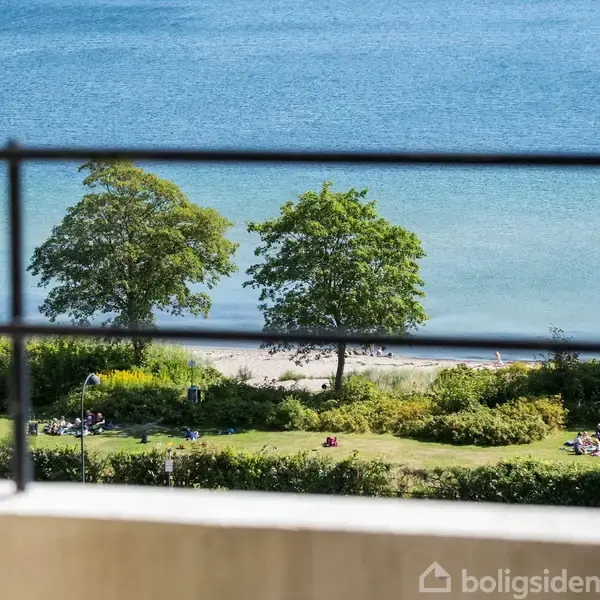 En balkon har udsigt over havet med klart blåt vand, omgivet af grønne træer og huse. En rund bordplade og blomsterpotte er placeret på altanen.