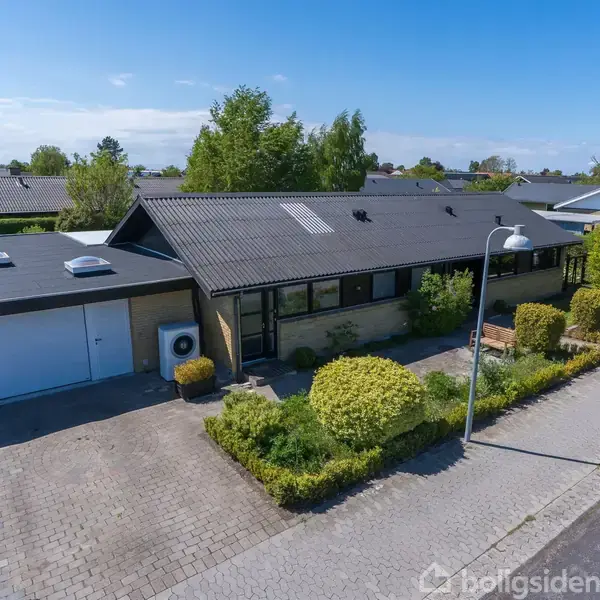Huset er et étplans parcelhus med fladt, sort tag, store vinduer og garage. Det ligger i et rækkehusområde med en flisebelagt indkørsel og omkranset af velplejet grøn beplantning.