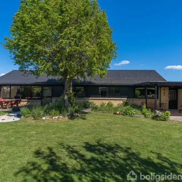 En lav, moderne bungalow med fladt, sort tag står i en grøn have. En stor træplæne omgiver huset med en centralt placeret træ, og en terrasse med havebord ses til venstre.