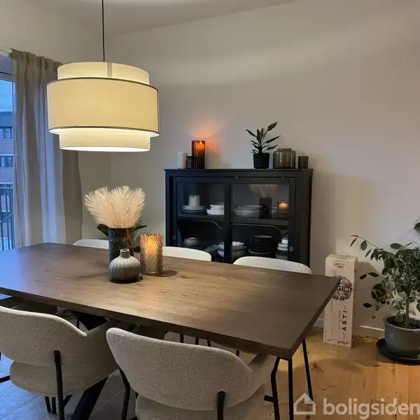 En spisestue med et træbord omgivet af stole. Bordet har vaser og stearinlys. En sort skænk med tallerkener står i baggrunden, og en lampe hænger over bordet.