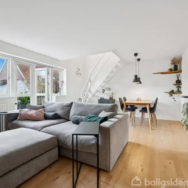 Grå sofa står i en stue ved et vindue med dagslys. Spisebord med stole ses i baggrunden nær en åbning til køkkenet. Indretning inkluderer planter og minimalistisk dekoration.