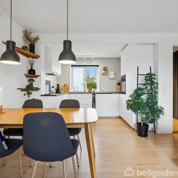 Spisebord med fire sorte stole står i et moderne køkken-alrum. To pendellamper hænger over bordet, mens grønne planter og lysestager tilføjer dekoration. Køkkenet har hvide skabe og en ovn.