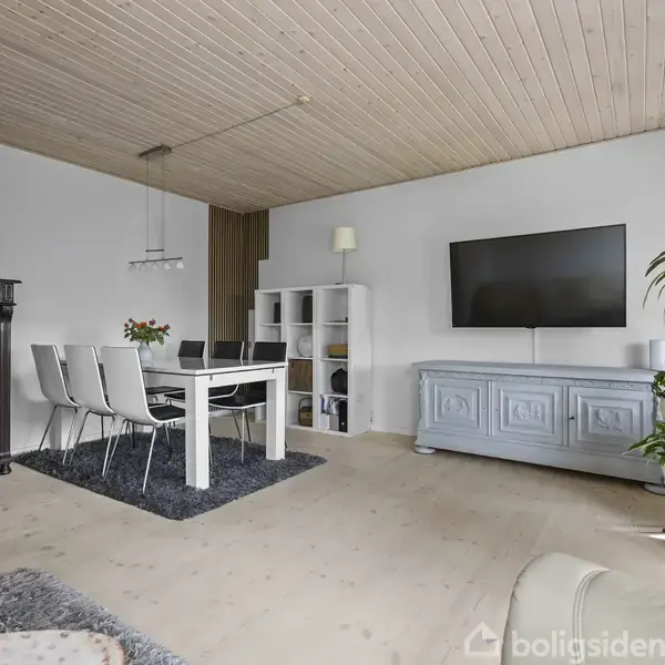 Et lyst stueområde med et spisebord og seks stole på et tæppe. Et skab og en reol står ved væggen. En stor fladskærm hænger over en blå kommode. Grønne planter ved vinduet.