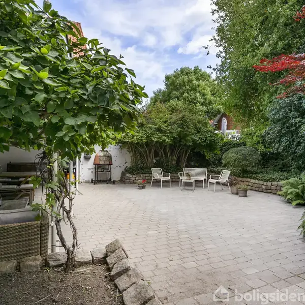 Terrasse med flisebelægning omgivet af frodig have. Rattanmøbler til venstre, grill i baggrunden og hvide havestole til højre skaber en indbydende udendørs opholdsplads.